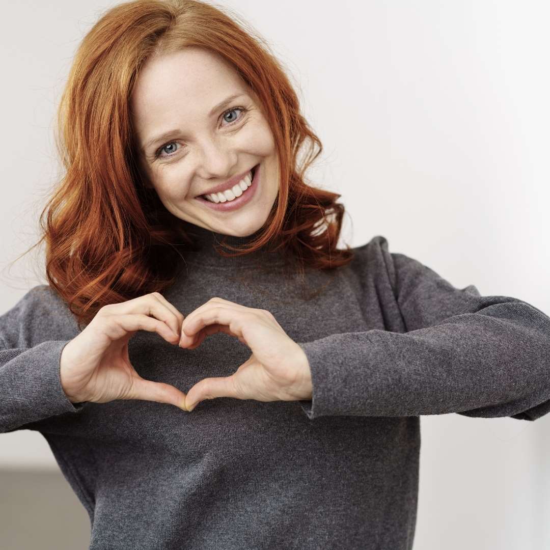 Rødhåret kvinde smiler til kameraet og former et hjerte med sine hænder.